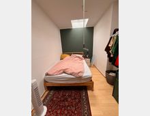 Schmales Schlafzimmer mit hellem Holzfußboden, einem roten Teppich und einem Holzbett mit rosa Bettwäsche. Eine grüne Wand mit Dachfenster befindet sich am Kopfende des Bettes; rechts steht ein Kleiderständer und eine Kommode, links ein weißer Standventilator.