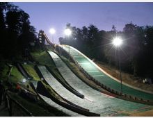 Eine beleuchtete Skisprungschanze am Abend, mit mehreren mit Kunststoffmatten bedeckten Sprung- und Landehügeln, umgeben von Bäumen im Hintergrund. Flutlichtmasten erhellen die Anlage, die von Wegen und Geländern eingefasst ist.