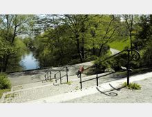 Steintreppe mit schmiedeeisernem Geländer führt hinunter zu einem Weg entlang eines Flusses, umgeben von dichtem Grün und Bäumen. Im Hintergrund ein geschwungener Weg und eine hölzerne Parkbank.