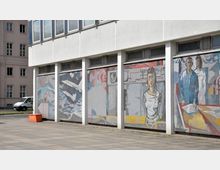 Flache Betonfassade eines Gebäudes mit großen Glasfenstern im oberen Bereich und bunten Wandmosaiken im unteren Bereich. Die Mosaike zeigen stilisierte Darstellungen von Personen, technischen Objekten und abstrakten Formen. Im Hintergrund ist ein historisches Gebäude zu sehen.