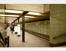 U-Bahnhof mit grün gekachelten Wänden, einer zentralen Säulenreihe und einem beleuchteten Informationsschild. Der Bahnsteig trägt das Schild „Neu-Westend“, im Hintergrund sind wartende Personen zu sehen.