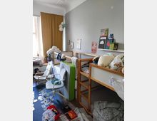 A children’s bedroom with wooden bunk beds against a gray wall, decorated with books, posters, and small wall art. The room features a window with mustard yellow curtains, a white cushioned chair, and scattered toys and books on a blue carpeted floor.