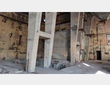 Ehemaliges Industriegebäude mit einer großen, offenen Halle, sichtbaren Backsteinwänden, Betonstützen und verrosteten Metallkonstruktionen. Der Boden ist stellenweise beschädigt, und offene Fenster sowie alte technische Installationen sind zu erkennen.