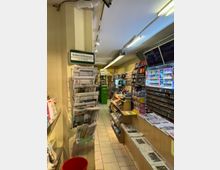 Kleiner Zeitungskiosk im Innenbereich mit Verkaufsregalen, die Zeitungen, Magazine und Tabakwaren präsentieren. Links befindet sich ein Zeitungsständer, rechts mehrere Verkaufsdisplays mit Kleinsüßwaren und ein Tresen mit ausliegenden Tageszeitungen.
