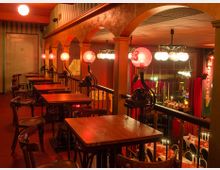 Ein gemütliches, zweistöckiges Restaurant mit warmer Beleuchtung, Holztischen und Stühlen. Im Obergeschoss befinden sich Tische entlang einer Geländerreihe mit dekorativen rosa Lampen, während der untere Bereich mit roten Vorhängen und gedeckten Tischen gestaltet ist.