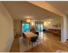 Ein modernes Wohnzimmer mit hellen Holzböden, einer großen Sitzecke und einem angrenzenden Essbereich mit einem Esstisch und Stühlen. Die rückseitige Wand besteht aus bodentiefen Fenstertüren, die auf einen Garten mit viel Grün blicken.