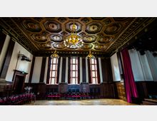 Historischer Saal mit reich verziertem Holzkassettendecke, großen verglasten Fenstern mit roten Rahmen und mehreren goldenen Kronleuchtern. An den Wänden sind holzvertäfelte Elemente sowie rote Vorhänge sichtbar, und Stühle sind entlang der Wand aufgereiht.