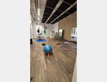 Large multipurpose studio room with wood flooring, a high ceiling with black acoustic panels and exposed pipes, and one long exposed brick wall. The space contains scattered exercise equipment such as a stability ball, cones, hoops, mats, and chairs, with windows and a bed-like platform along one side.