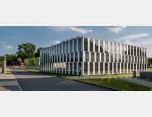 Modernes Bürogebäude mit einer Fassade aus großen Glas- und Metallpaneelen, umgeben von Grünflächen und Gehwegen. Im Hintergrund sind Bäume und ein traditionelles Fachwerkhaus zu sehen.