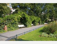 Ein gepflegter Parkweg mit Pflastersteinen, gesäumt von weiß gestrichenen Bänken und dicht bewachsenen Rankgittern. Auf der rechten Seite befinden sich gepflegte Blumenbeete und eine Wiese; im Hintergrund sind Bäume und ein mehrstöckiges Gebäude zu sehen.