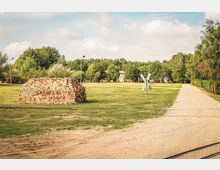 Eine ländliche Szenerie mit einem Stapel Ziegelsteinen auf einer Wiese, einem Schotterweg, Bahngleisen im Vordergrund und einem Verkehrsschild in Kreuzform. Im Hintergrund sind Bäume, eine kleine Gebäudeansammlung und ein bewölkter Himmel zu sehen.