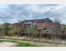 Backsteingebäude mit mehreren Etagen und großen Fenstern, umgeben von Bäumen und Büschen, im Vordergrund ein kleiner Wasserlauf und eine Wiese; der Himmel ist wolkenbedeckt.
