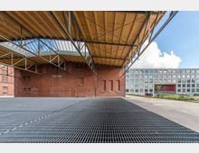 Freistehende Überdachung mit Holzdach und Metallrahmen über einem Gitterboden, vor einer roten Ziegelwand eines Gebäudes; im Hintergrund ein modernes Gebäude mit rechteckigen Fenstern und einem roten Durchgang.