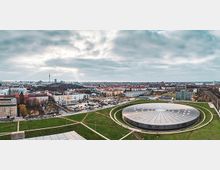 Panoramablick über eine weitläufige Stadtlandschaft mit dicht bebauten Wohn- und Gewerbegebieten unter bewölktem Himmel. Im Vordergrund liegt eine große, runde Arena mit flachem, radial gegliedertem Dach, umgeben von Grünflächen und Wegen.