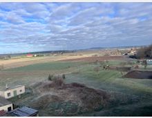 Ländliche Landschaft mit Feldern, vereinzelt stehenden Bäumen und Weiden. Im Hintergrund befinden sich Windräder, Wälder und verstreute Gehöfte unter einem teils bewölkten Himmel.