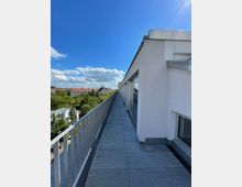 Ein langer Balkon auf einem modernen Gebäude mit grauen Bodenfliesen und einem Metallgeländer auf der linken Seite. Rechts befinden sich Fensterfronten, im Hintergrund sind eine Baumlandschaft und Dächer weiterer Gebäude zu sehen, unter einem klaren blauen Himmel mit Wolken.