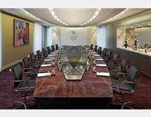 Konferenzraum mit einem langen, ovalen Holztisch, umgeben von schwarzen Bürostühlen. Der Raum ist modern gestaltet, mit einer hinteren Wand, die das Logo "Waldorf Astoria Berlin" trägt, und einer seitlichen Abstellfläche mit Marmorverkleidung und Dekorationselementen.