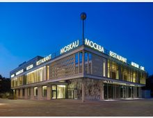 Modernes zweistöckiges Gebäude bei Dämmerung mit großflächigen Glasfronten und geometrischen Fassadenelementen. Auf dem Dach leuchten Schriftzüge wie "Moskau" und "Restaurant", während an der Seite ein Wandmosaik zu sehen ist.