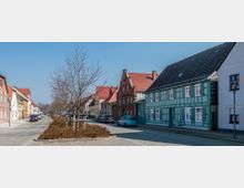 Straßenansicht in einer Kleinstadt mit Kopfsteinpflaster, links und rechts gesäumt von Reihenhäusern. Auffällig ist ein grünes Fachwerkhaus und ein rotes Backsteingebäude, sowie kahle Bäume und parkende Autos entlang der Straße.