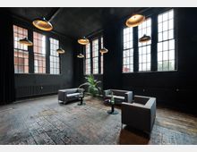 Industriell gestalteter Raum mit hohen Decken, dunklen Wänden und großen Fenstern mit Gitterrahmen. In der Mitte stehen vier graue Sessel, zwei runde Beistelltische mit Dekorationen und eine Zimmerpflanze auf einem rustikalen Holzboden.