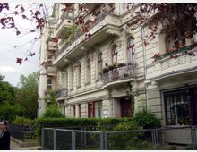Mehrstöckiges historisches Wohngebäude mit verzierten Fassaden, schmiedeeisernen Balkongeländern und roten Fensterrahmen. Im Vordergrund befindet sich ein kleiner Garten mit Sträuchern, umgeben von einem Metallzaun.