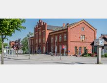 Historisches Bahnhofsgebäude aus rotem Backstein mit verzierten Zierelementen und großen Fenstern. Im Vordergrund sind Fahrradständer, eine Bushaltestelle, eine Uhr auf einer Säule und ein Fußweg zu sehen, umgeben von vereinzelten Bäumen und Schildern.