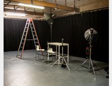 Fotostudio mit grauem Boden, schwarzem Vorhanghintergrund und Metallträgern an der Decke. Im Vordergrund stehen ein Klapptisch mit zwei weißen Stühlen, eine Aluminiumleiter sowie professionelle Beleuchtungsausrüstung auf Stativen.