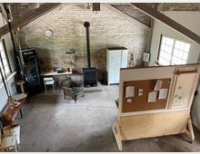 Industrieller Arbeitsraum mit Sichtziegelwänden, sichtbaren Holzträgern und Betonboden. Im Raum befinden sich ein schwarzer Kamin, ein Kühlschrank, ein Schreibtisch mit Schreibmaschine, ein Sessel, eine Pinnwand sowie große Fenster, die Tageslicht hereinlassen.