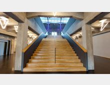 Breiter Treppenaufgang aus Holz mit schwarzen Seitengeländern in einem Gebäude mit Betonwänden und modernen, leuchtenden Lampen in geometrischen Formen. Im Hintergrund große Fenster mit Sicht auf eine Ziegelwand und eine hohe Decke.