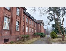 Backsteinbau mit großen Fenstern im Stil der klassischen Architektur, flankiert von einem schmalen Gehweg und vereinzelten Bäumen und Sträuchern. Die Umgebung wirkt leicht verwildert, mit kahlen Ästen und ungemähtem Gras.