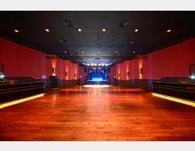 Ein leerer Veranstaltungsraum mit Holzboden, roten Wänden und einer schwarzen Decke mit eingelassenen Deckenleuchten. Am Ende des Raumes befindet sich eine Bühne, die mit bunten Scheinwerfern beleuchtet ist.