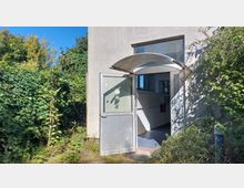 Eine geöffnete graue Metalltür mit Glaseinsatz, die in ein schlichtes Gebäude führt. Im Vordergrund sind dichte Büsche und Pflanzen zu sehen, im Hintergrund ein heller Innenraum mit weißen Wänden und Feuermeldern.