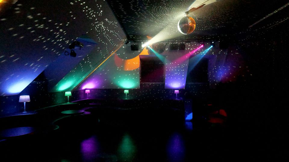 Innenraum eines Nachtclubs mit schräger Decke und einer Discokugel, die Lichtpunkte über Wände und Decke streut. Bunte Scheinwerfer und indirekte Beleuchtung erhellen eine dunkle Tanzfläche mit niedrigen Sitzbereichen und kleinen Tischlampen entlang der Seiten.