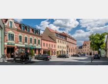 Städtischer Platz mit historischen Gebäuden, darunter ein Hotel, ein Restaurant und weitere Geschäfte, die farbige Fassaden mit klassischen Fensterrahmen und roten Ziegeldächern aufweisen. Im Vordergrund stehen Autos geparkt, ein Baum spendet Schatten, und der Himmel ist leicht bewölkt.