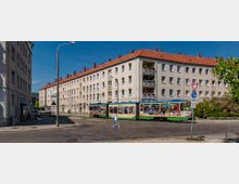 Straßenansicht mit einer beige verputzten Wohnhausreihe mit roten Ziegeldächern und Balkonen, davor eine bunte Straßenbahn auf einer gepflasterten Straße. Im Vordergrund ein Gehweg mit einem Fußgänger und Verkehrsschildern, im Hintergrund vereinzelte Bäume.