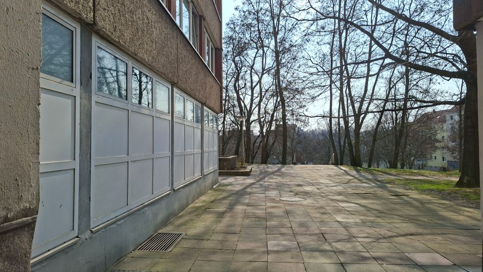 Gepflasterter Gehweg neben einem mehrstöckigen Wohngebäude mit großen verglasten Balkon- oder Loggiaelementen an der Fassade. Rechts stehen kahle Bäume auf einer Grünfläche, im Hintergrund sind weitere Wohnhäuser zu sehen.