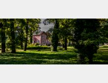 Ein rosa-rotes Gebäude im Stil der alten Architektur mit weißen Fensterrahmen und einem Satteldach, das inmitten eines Parks mit hohen, dicht bewachsenen Bäumen und einer offenen Rasenfläche steht. Im Vordergrund befinden sich Schatten der Bäume und grünes Blattwerk.