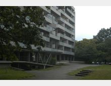 Ein mehrstöckiges Wohngebäude mit Balkonen und einer angrenzenden Grünfläche. Im Vordergrund führt ein gepflasterter Weg vorbei, umgeben von Bäumen und Wiese.