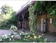 Ein ländlicher Innenhof mit Kopfsteinpflaster, flankiert von einem Backsteingebäude mit weißen Türen und grünen Fensterläden. Rankende Pflanzen und Blumenrabatten mit weißen, gelben und rosa Blüten umgeben den Bereich, während ein hölzernes Vordach mit hängenden Pflanzen für Schatten sorgt.