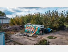 Container auf einem verlassenen Gelände mit einer bunten Wandbemalung, die einen geometrischen Fuchs in lebhaften Farben darstellt, umgeben von Bäumen und wildem Gestrüpp. Daneben stehen Mülltonnen, ein Stapel Steine und Gartenliegen unter einer bewölkten Himmelsszenerie.