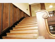 Holztreppe in einem historischen Gebäude mit kunstvoll geschnitztem Handlauf, holzverkleideten Wänden und einer Decke mit verzierten Stuckelementen. Im Hintergrund ist ein Kronleuchter sowie ein Geländer im oberen Stockwerk zu sehen.