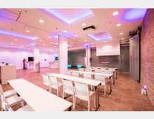 Moderner Veranstaltungsraum mit weißen Tischen und Stühlen in Reihen angeordnet, einer Präsentationsfläche mit Beamerbild auf einer Leinwand und einer Backsteinwand an der rechten Seite. Die Decke ist mit farbiger Beleuchtung und eingelassenen Leuchten gestaltet.