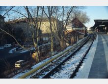 Ein verschneiter Bahnsteig mit Gleisen, flankiert von Bäumen, die eine nahe gelegene Straße und Gebäude mit traditionellen Ziegeldächern und Wohnhausfassaden im Hintergrund einrahmen. Der Bahnsteig hat eine Überdachung auf der rechten Seite.