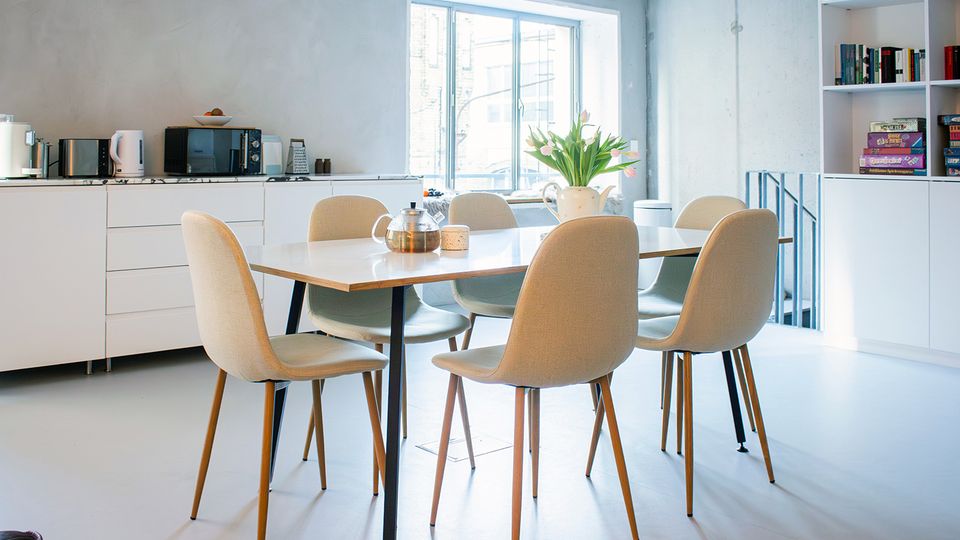 Heller, moderner Küchen- und Essbereich mit großem Fenster, weißer Küchenzeile mit Elektrogeräten und einem Esstisch mit sechs gepolsterten Stühlen. Rechts steht ein offenes Regal mit Büchern und Spielen; auf dem Tisch stehen Teekanne und eine Vase mit Blumen.