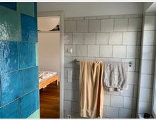 Badezimmer mit hellen weißen Fliesen an der Wand und blauen glänzenden Kacheln auf der linken Seite. Ein Handtuchhalter mit einem gelben und einem gemusterten Handtuch ist sichtbar, rechts befindet sich ein Fenster, und im Hintergrund ist ein Holzbett in einem angrenzenden Raum erkennbar.