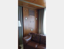 Innenraum mit einer Wand aus Holzvertäfelung, die runde dekorative Ausschnitte und ein Lüftungsgitter zeigt. Im Vordergrund befinden sich ein braunes Ledersofa, eine Stehlampe und seitlich lange Vorhänge vor einem Fenster.