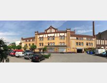 Eine historische Industriegebäude-Fassade aus gelbem Klinker mit roten Backsteinverzierungen, Rundbogenfenstern und einem dekorativen Zinnenabschluss. Vor dem Gebäude befindet sich ein gepflasterter Hof mit geparkten Autos, Bäumen und einem Schornstein im Hintergrund.