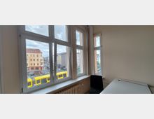 Heller Büroraum mit großen, weiß gerahmten Fenstern und Blick auf eine städtische Kreuzung mit mehrstöckigen Gebäuden und einer gelben Straßenbahn. Im Inneren sind eine weiße Tischplatte, ein schwarzer Stuhl und ein Heizkörper unter der Fensterbank zu sehen.