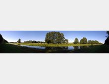 Panorama einer Flusslandschaft mit ruhigem Wasser, das Bäume und Wiesen am Ufer spiegelt. Im Hintergrund sind weitläufige grüne Felder, vereinzelt Bäume und ein kleines Gebäude sichtbar, unter einem klaren blauen Himmel.