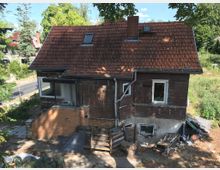 Ein älteres Wohnhaus mit Holzwänden und rotem Ziegeldach, teilweise renovierungsbedürftig. Im Vordergrund befindet sich ein kleiner Hof mit gestapelten Bau- und Abfallmaterialien, umgeben von grüner Vegetation und einem asphaltierten Gehweg.